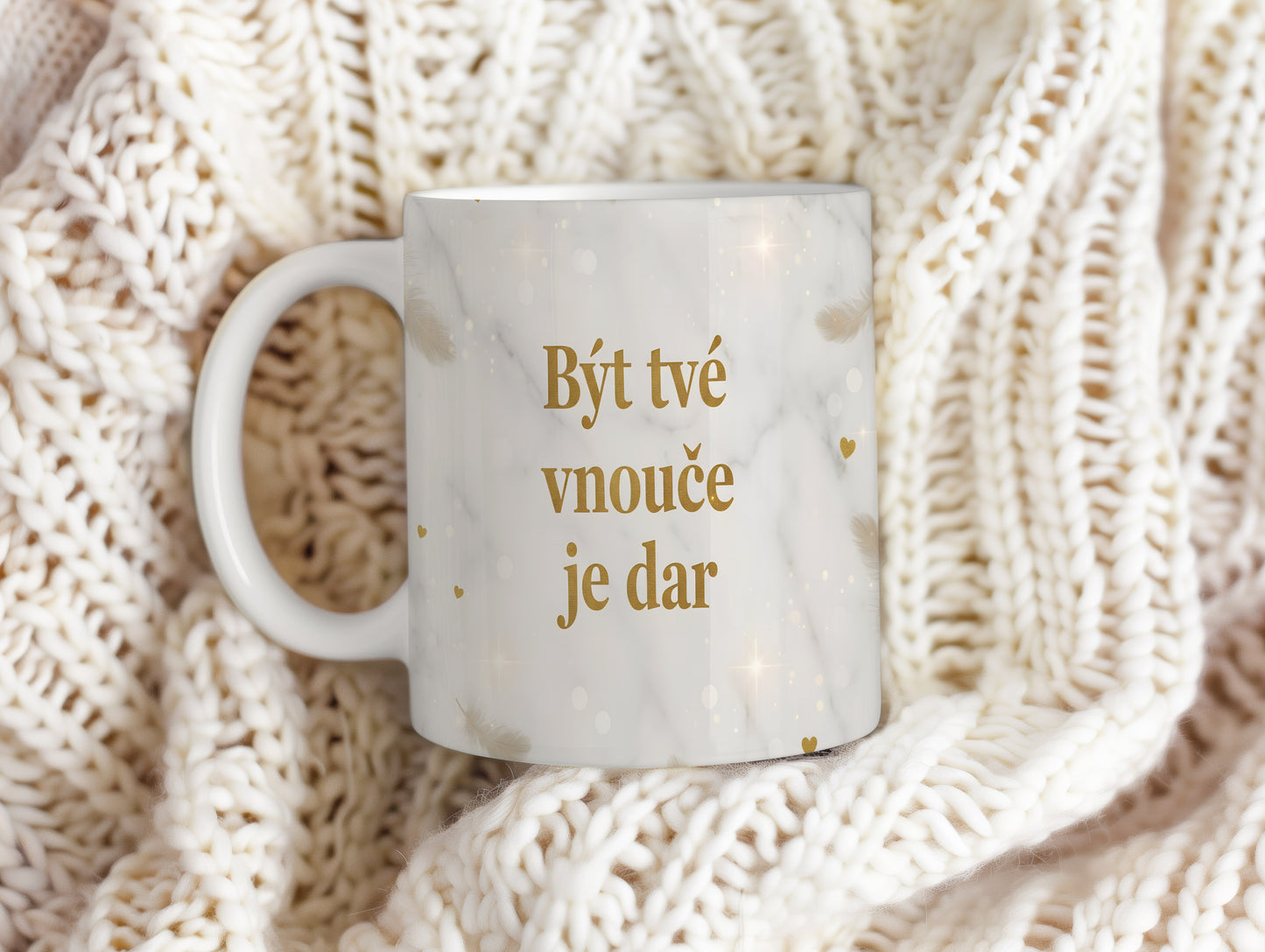 Hrnek s nápisem 'Být tvé vnouče je dar' se zlatými peříčky na mramorovém pozadí.