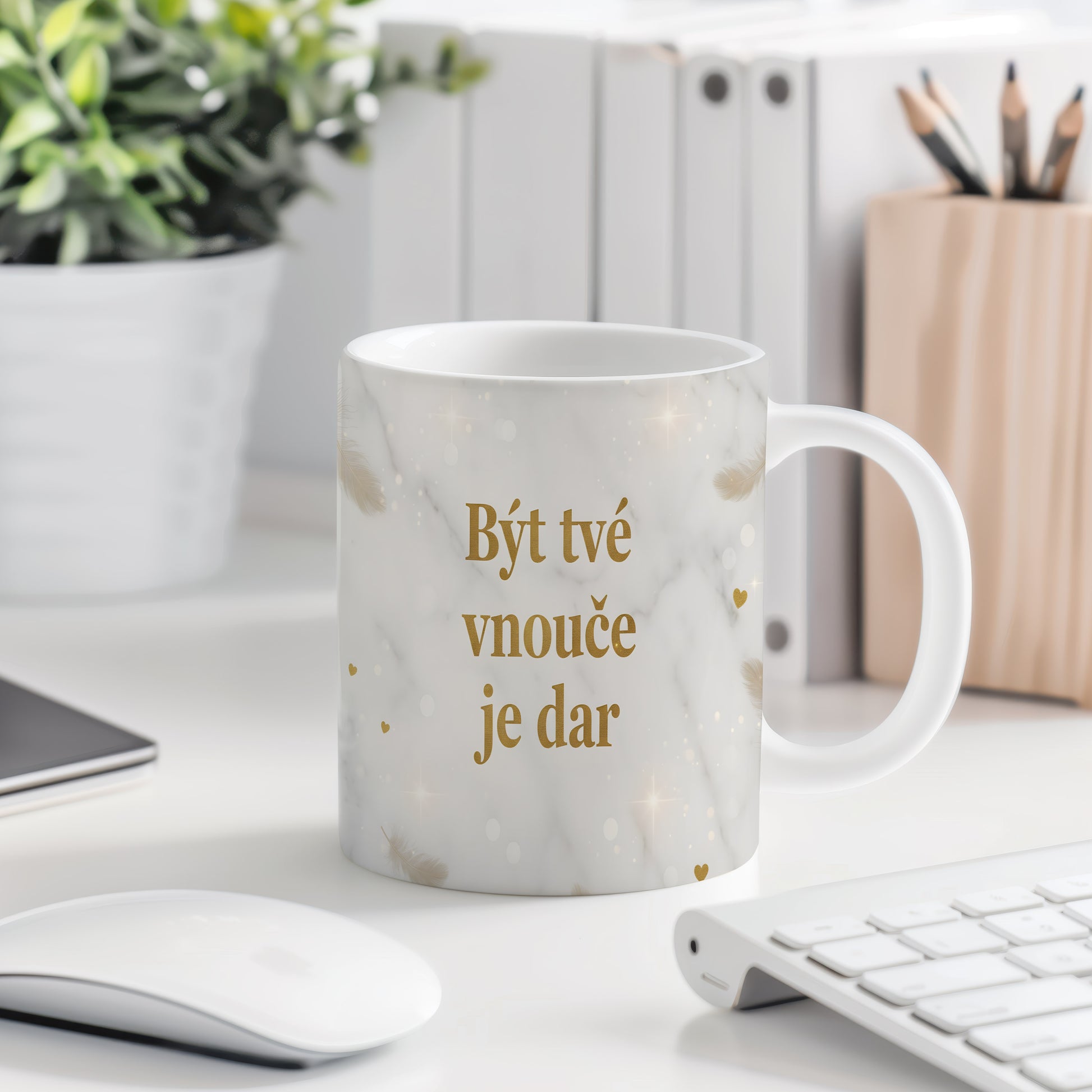 Hrnek s nápisem 'Být tvé vnouče je dar' se zlatými peříčky na mramorovém pozadí.
