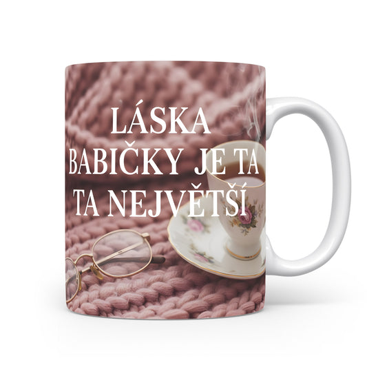Hrnek s nápisem LÁSKA BABIČKY JE TA NEJVĚTŠÍ na růžovém pleteném pozadí.