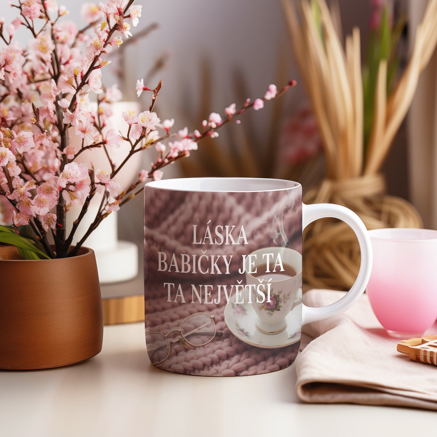 Hrnek s nápisem LÁSKA BABIČKY JE TA NEJVĚTŠÍ na růžovém pleteném pozadí.