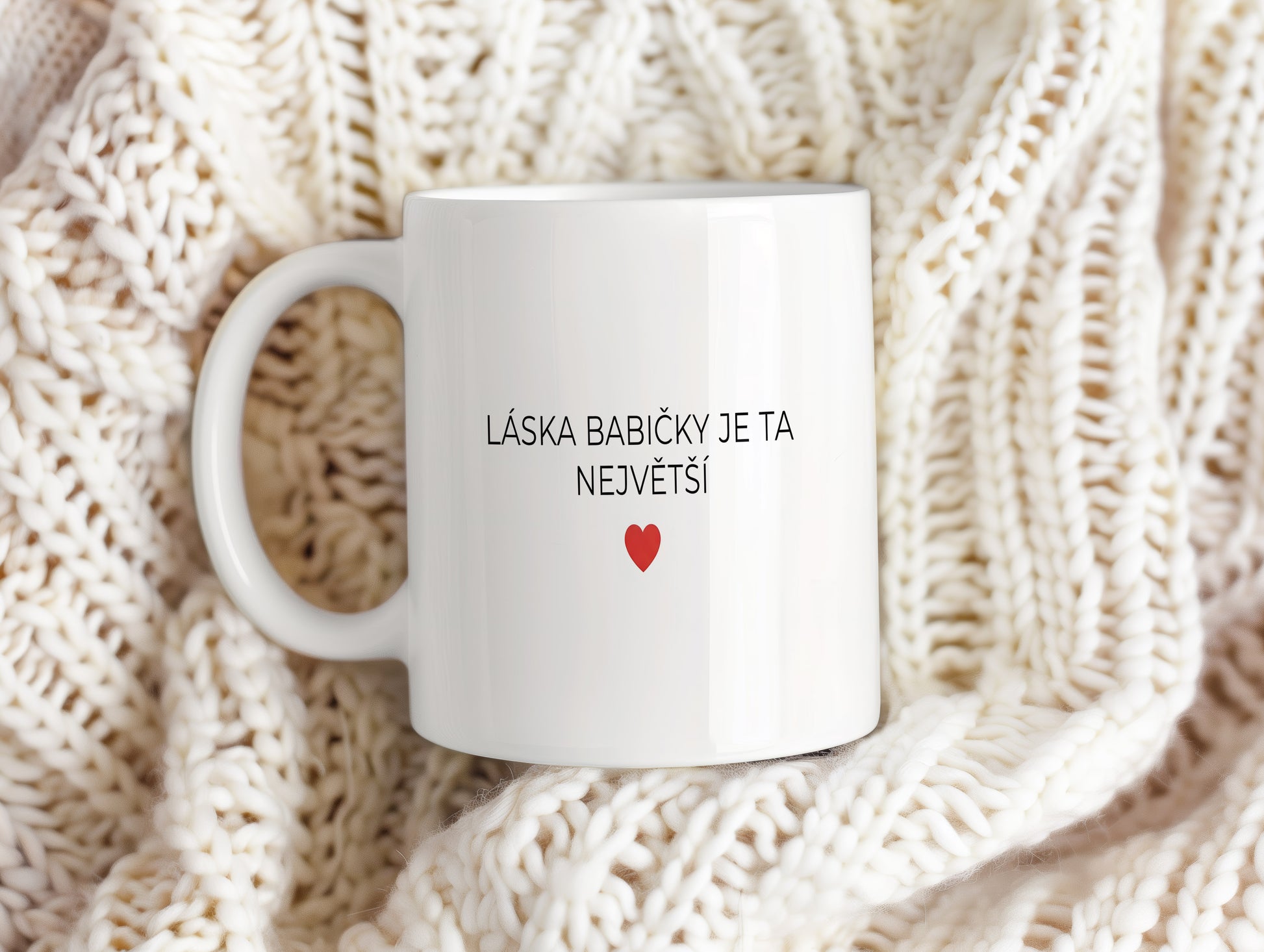 Bílý hrnek s nápisem 'LÁSKA BABIČKY JE TA NEJVĚTŠÍ' a červeným srdcem.