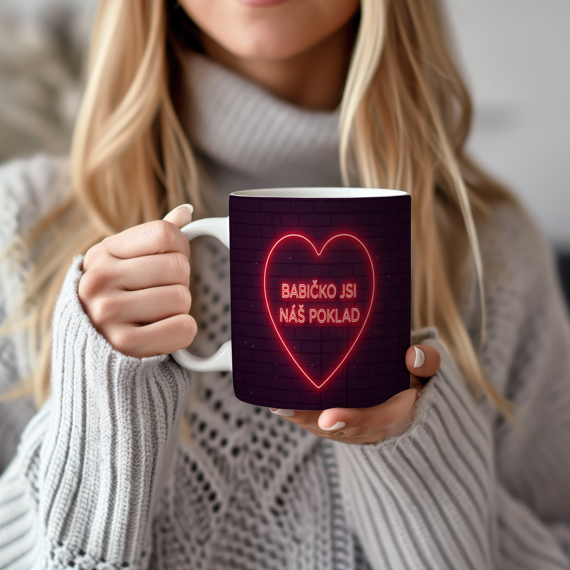 Hrnek s neonovým srdcem a nápisem BABIČKO JSI NÁŠ POKLAD na cihlovém pozadí.