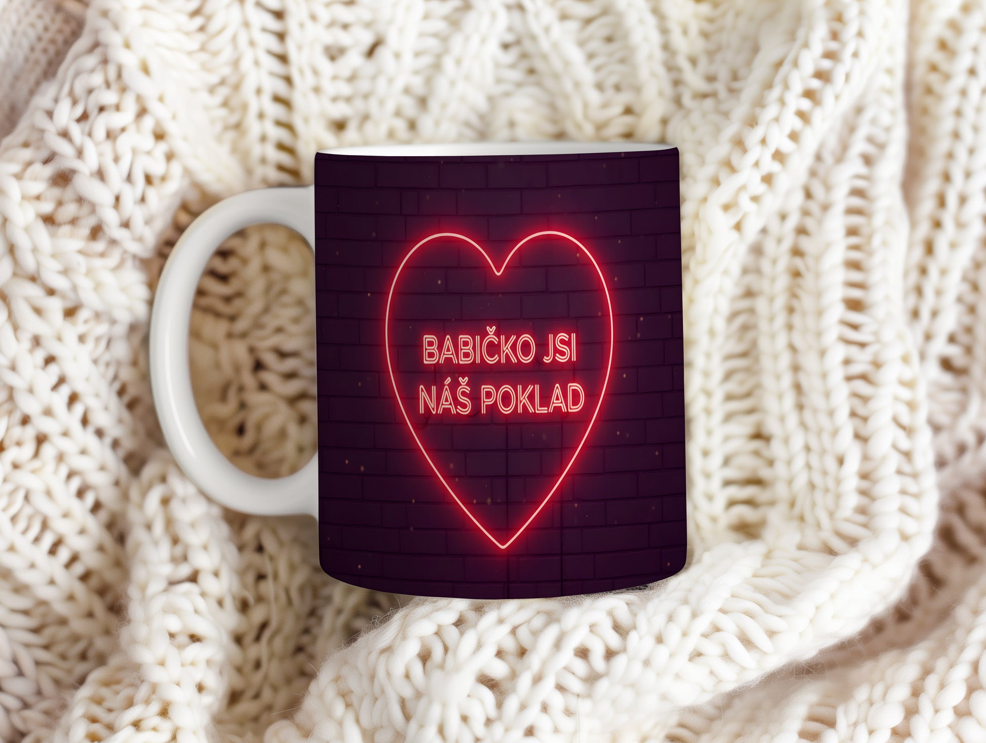 Hrnek s neonovým srdcem a nápisem BABIČKO JSI NÁŠ POKLAD na cihlovém pozadí.