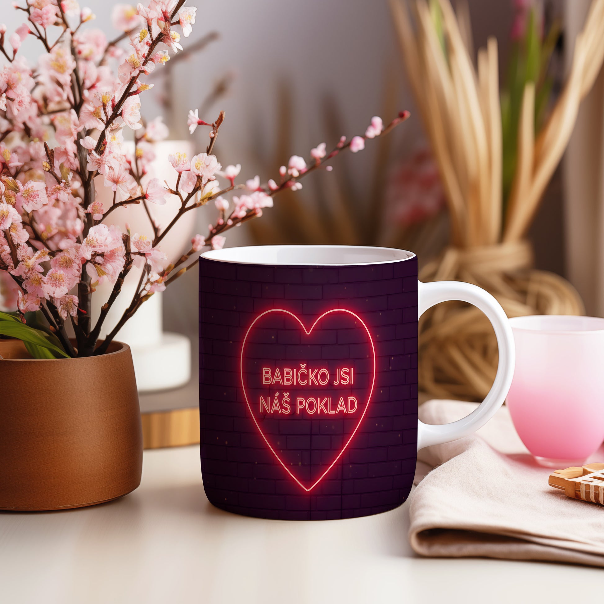 Hrnek s neonovým srdcem a nápisem BABIČKO JSI NÁŠ POKLAD na cihlovém pozadí.