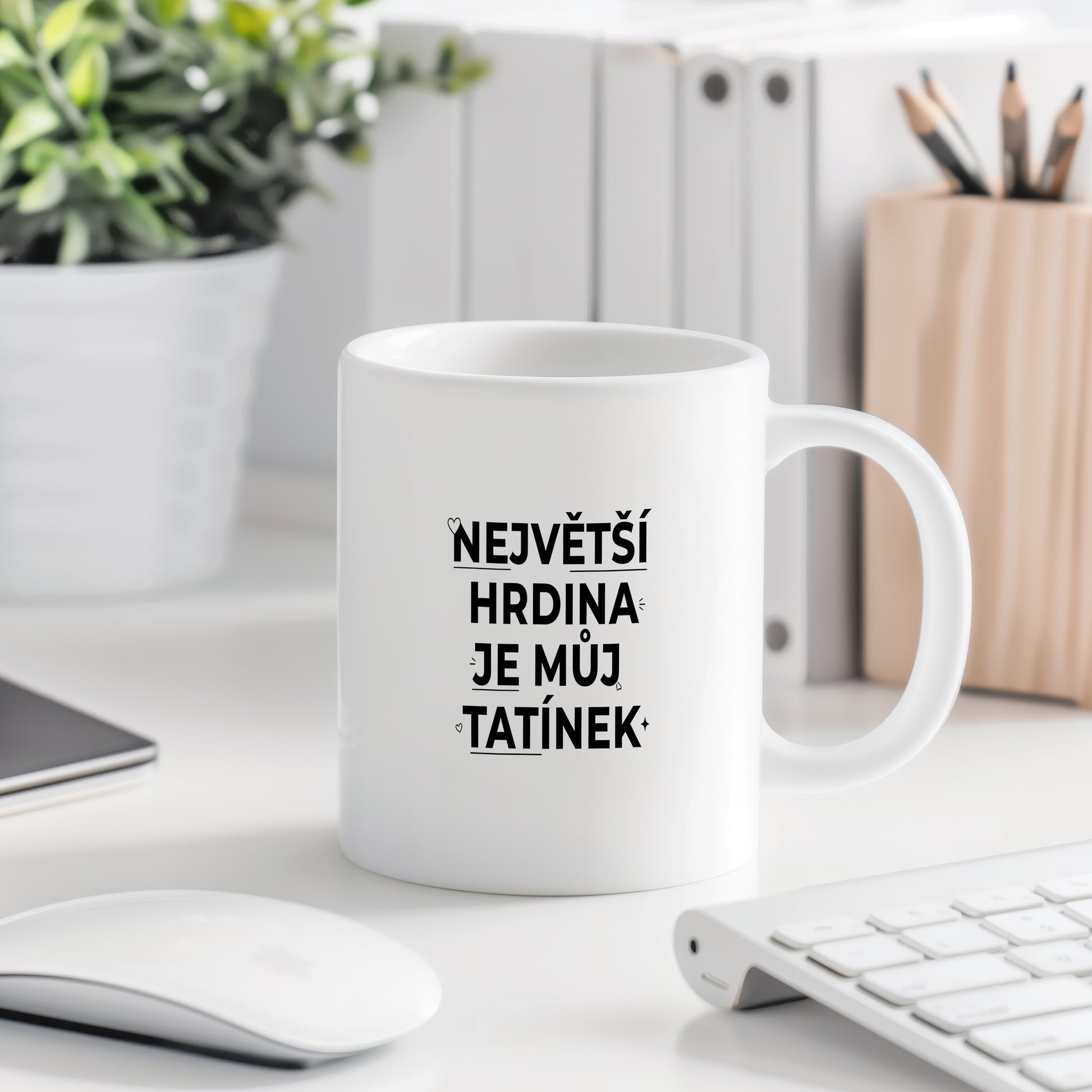 Bílý hrnek s černým nápisem 'NEJVĚTŠÍ HRDINA JE MŮJ TATÍNEK' a malými srdíčky.