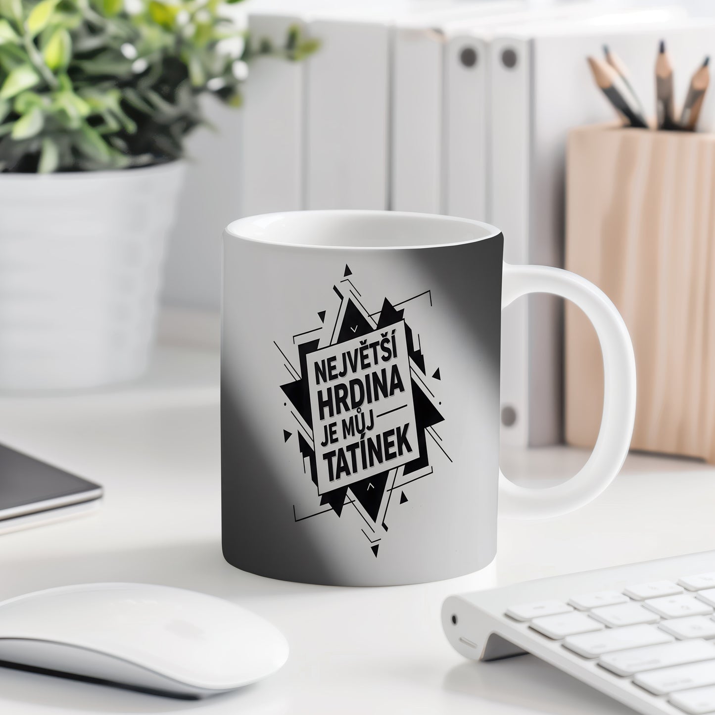 Bílý hrnek s černým nápisem 'NEJVĚTŠÍ HRDINA JE MŮJ TATÍNEK' a geometrickým vzorem.