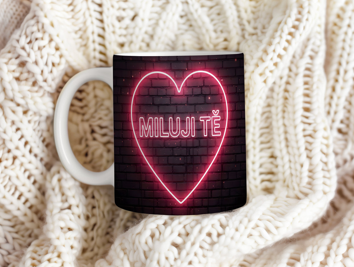 Hrnek s neonovým nápisem 'MILUJI TĚ' na cihlovém pozadí.