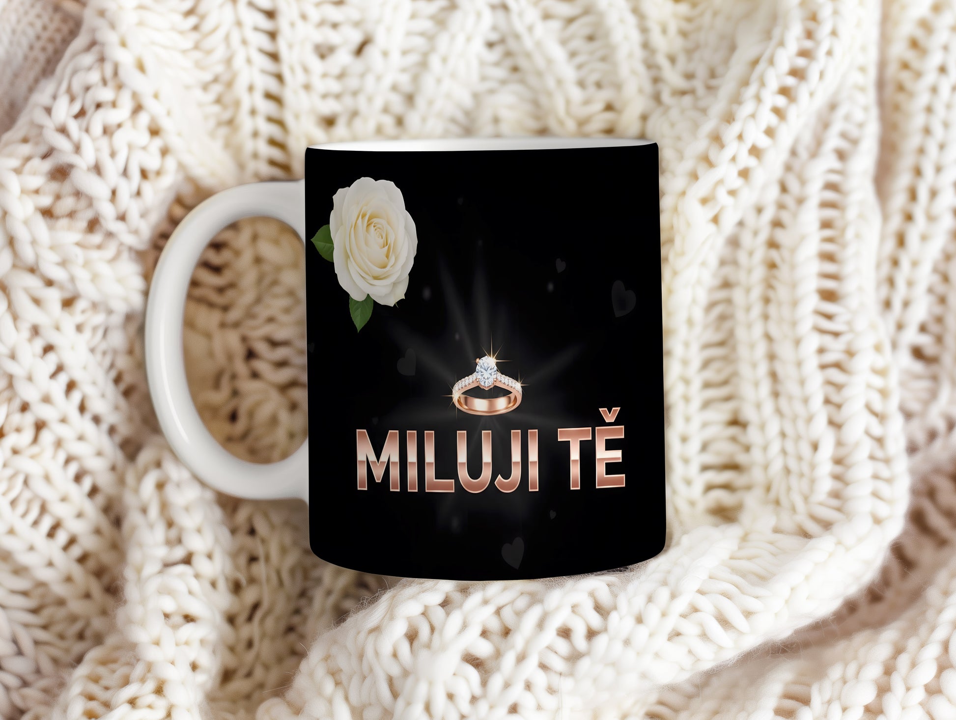 Černý hrnek s nápisem MILUJI TĚ, prstenem a bílou růží.