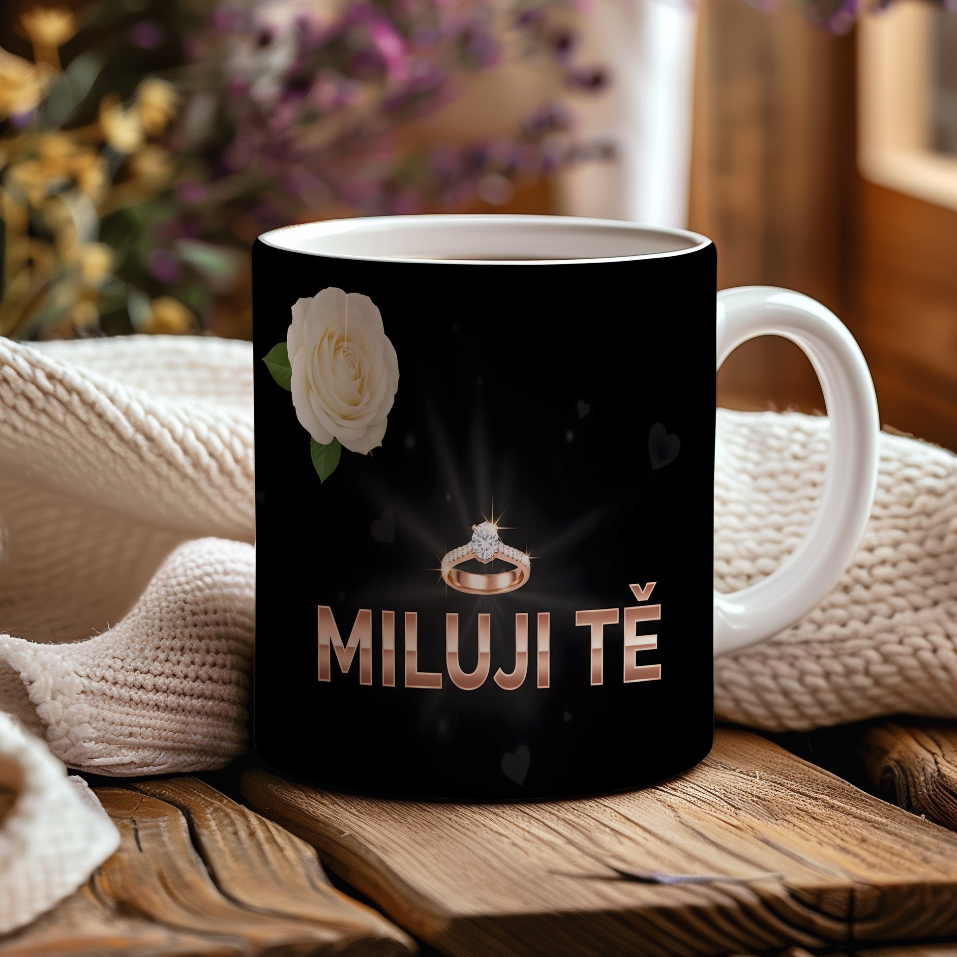 Černý hrnek s nápisem MILUJI TĚ, prstenem a bílou růží.