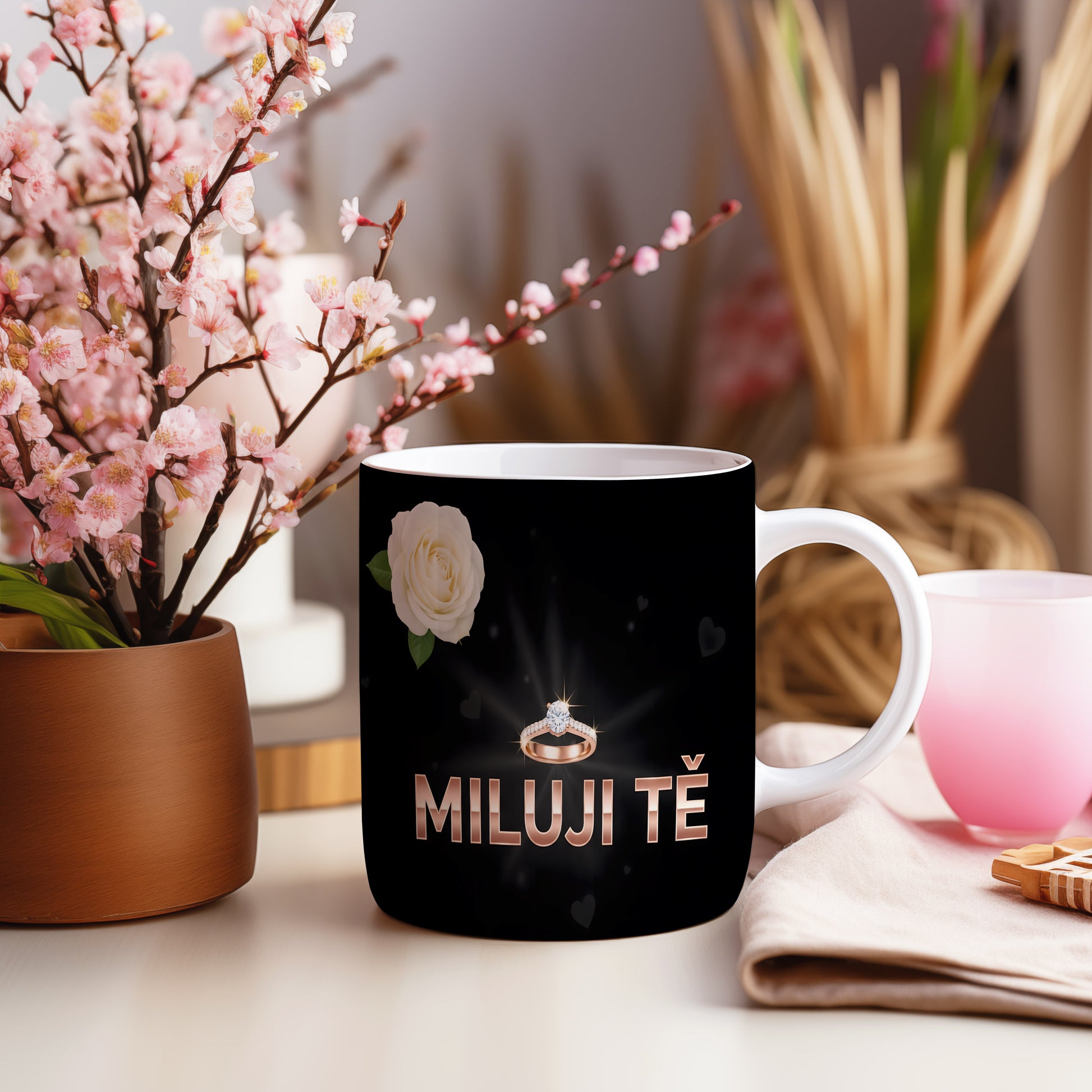 Černý hrnek s nápisem MILUJI TĚ, prstenem a bílou růží.
