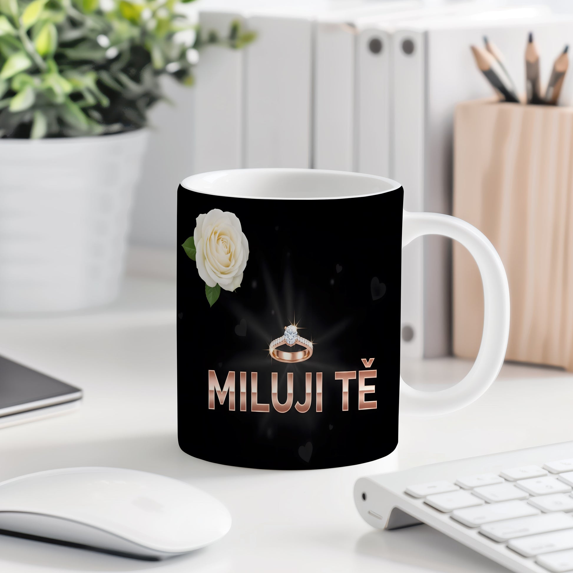 Černý hrnek s nápisem MILUJI TĚ, prstenem a bílou růží.